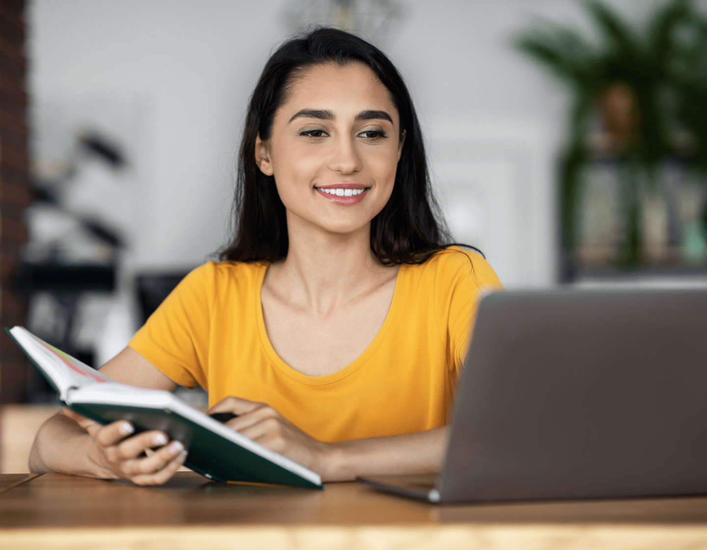 An image of a woman looking at her laptop on a wooden bench. She has a notebook in her right hand and a pen in her left hand